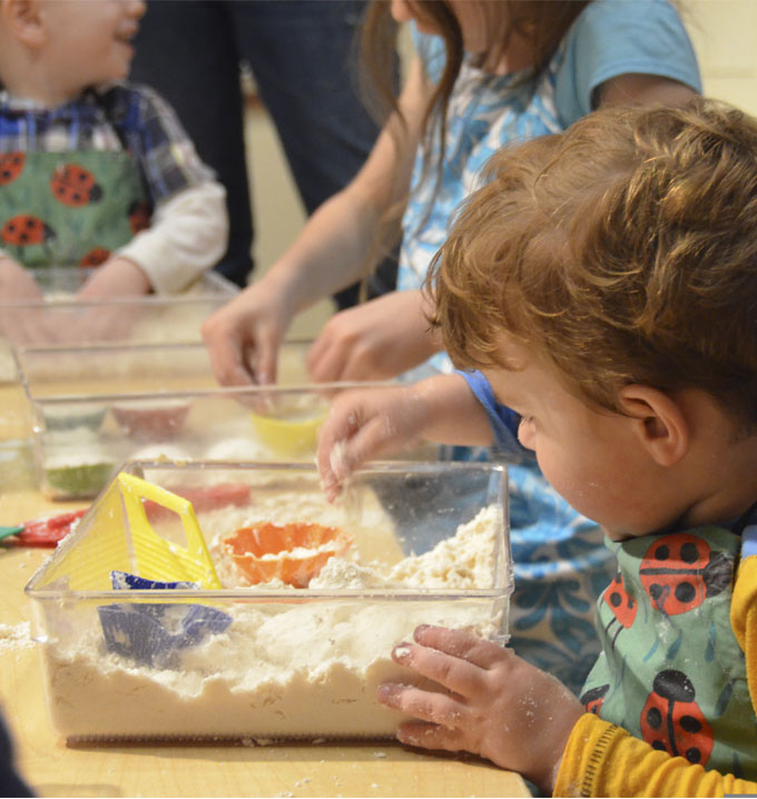 Three children stood around a table with their hands in boxes of flour wearing ladybug aprons.