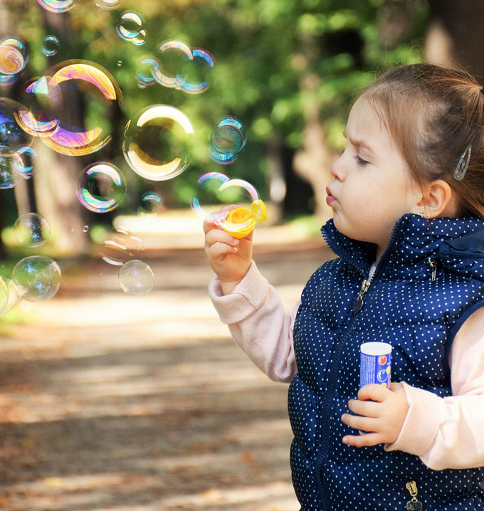 A child in a blue gilet with brown hair stood in a wooded area blowing bubbles.