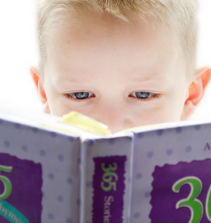 A close-up shot of a child with blonde hair and blue eyes reading a purple storybook close-up against his face.