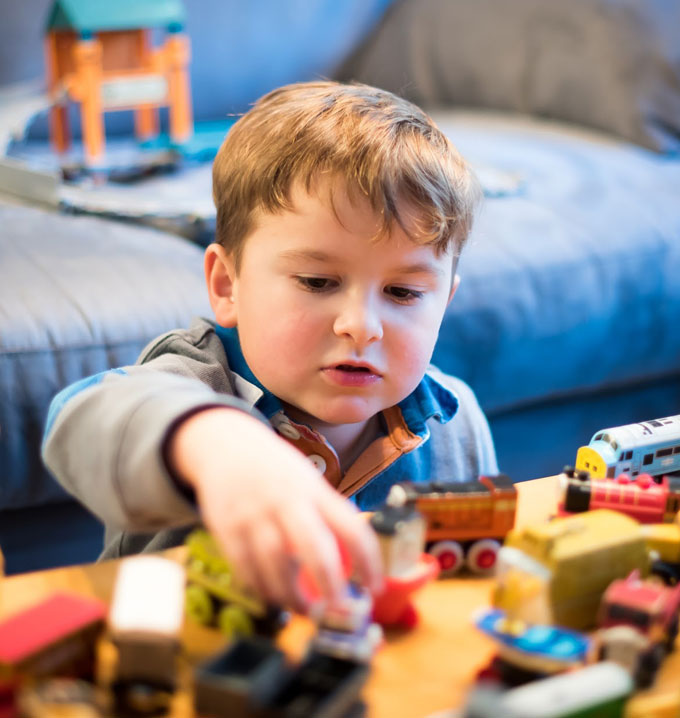 A child sat on the floor facing a wooden table playing with lots of wooden trains in front of a blue couch.