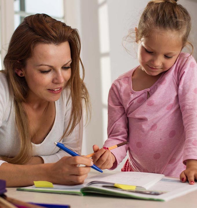 A woman with brown long hair and a white shirt leaning over a table holding a blue pen next to a girl wearing pink smiling holding a pen.