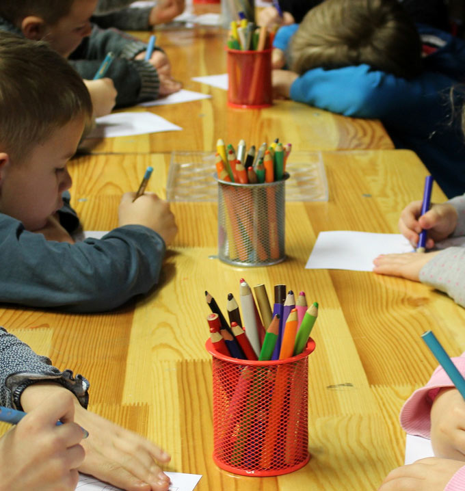 Children sat at a table opposite each other with their heads leaning on tables drawing in front of three pots of coloring pencils.