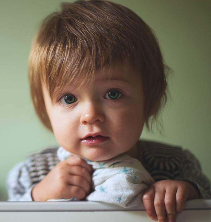 A child with blonde short hair and green eyes holding a small blanket in one hand in a cot.