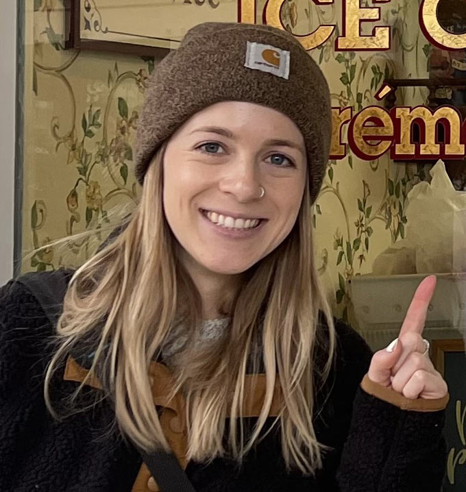 A woman with long blond hair wearing a brown beanie and black coat smiling and pointing at a window.