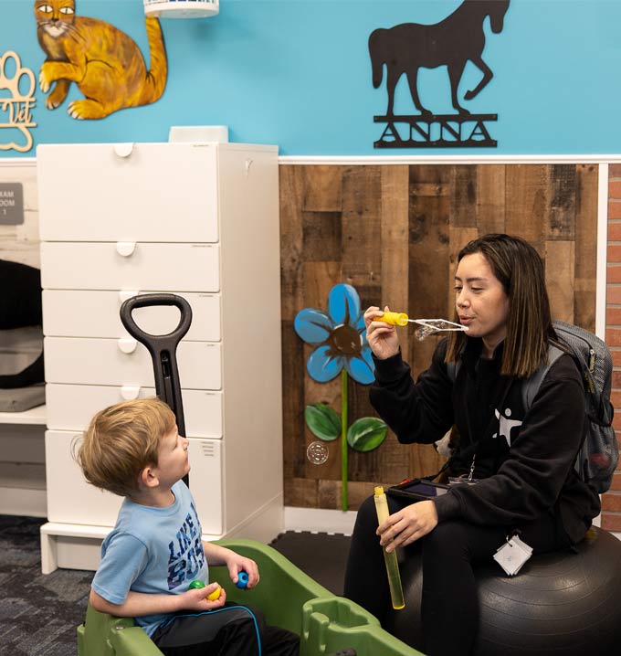 A Lighthouse Autism Center staff member in black uniform sat on an exercise ball blowing bubbles towards a child sat in a toy car.