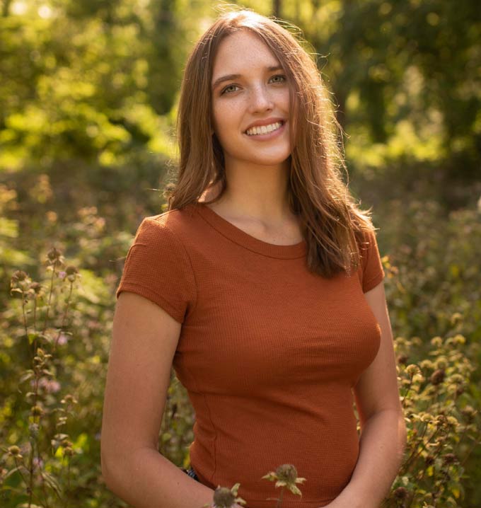 A brunette woman in a meadow wearing a brown t-shirt, smiling at the camera.