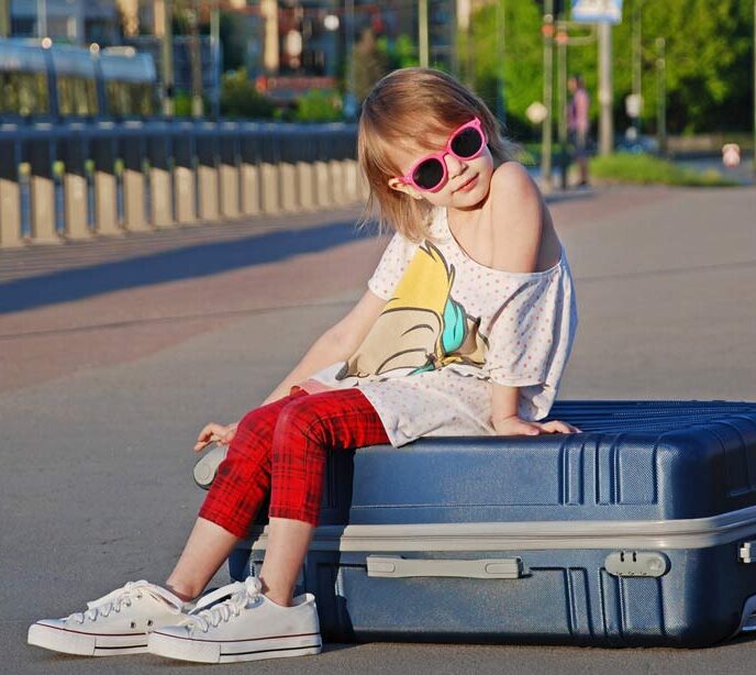 A child wearing adult sized sneakers and pink sunglasses sat on top of a blue suitcase