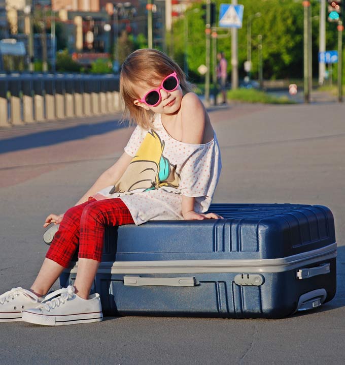 A child with blonde hair wearing pink sunglasses, red trousers and a white shirt sat on top of a large blue piece of luggage.