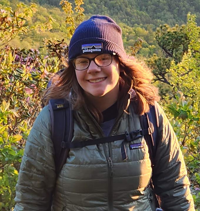 A woman wearing a gray puffer jacket, beanie hat and glasses in front of green bushes.