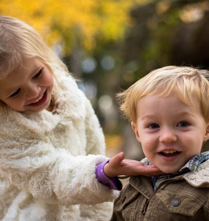 A girl in a white jacket smiling looking down and holding her hand on another child's chin smiling.