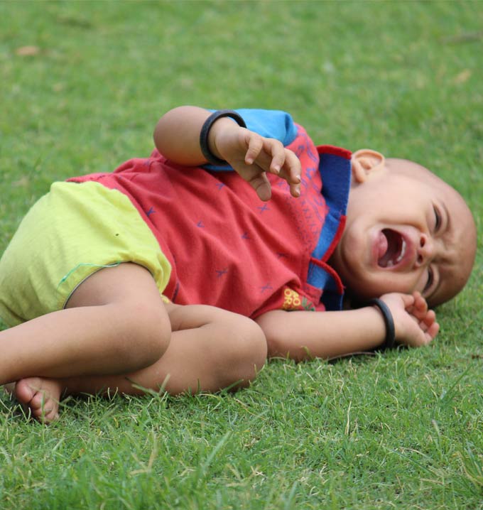 A child wearing a red shirt and yellow shorts lying down on the grass crying and having a tantrum.