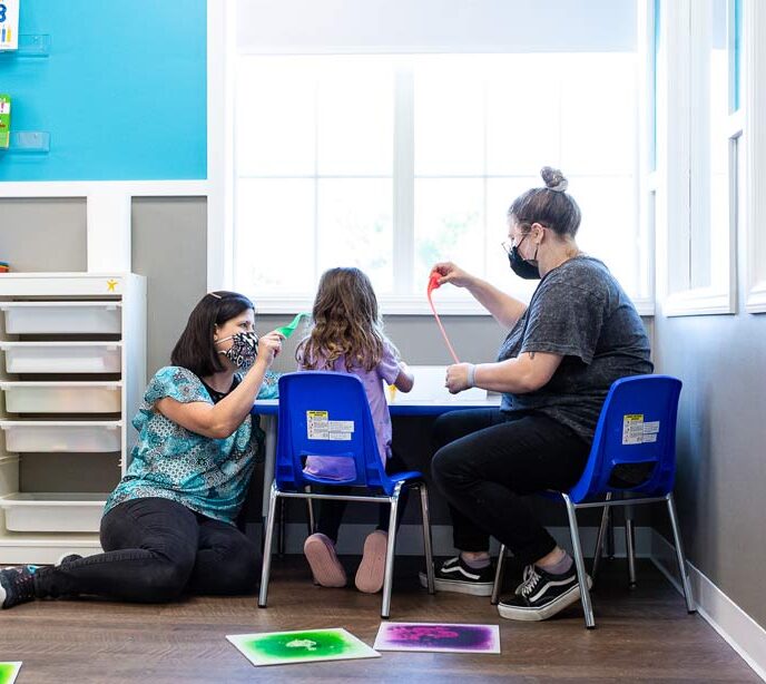 Two ladies wearing face masks sat at a table with a child playing with putty