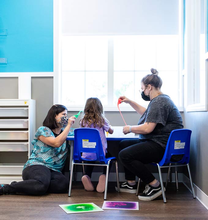 Two Lighthouse Autism Center members of staff sat at a table wearing face masks playing with slime with a child