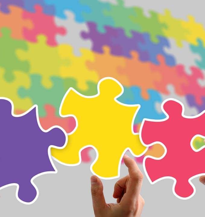 Multiple colored puzzle pieces in the background and a hand holding a yellow puzzle piece in the center of two others.