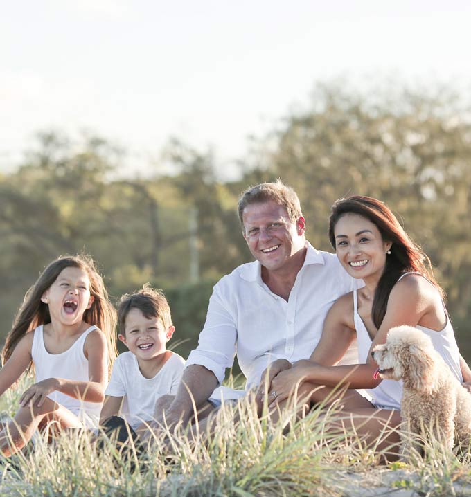 A family of a mom, dad, a female child and male child sat in a row all wearing white clothing with their dog in a field similing.