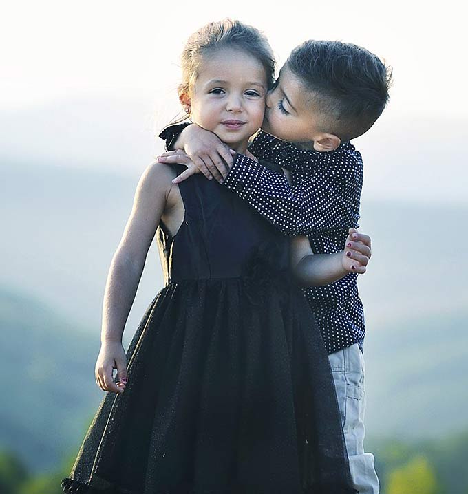 A boy with his arms around a girl and kissing her cheek.