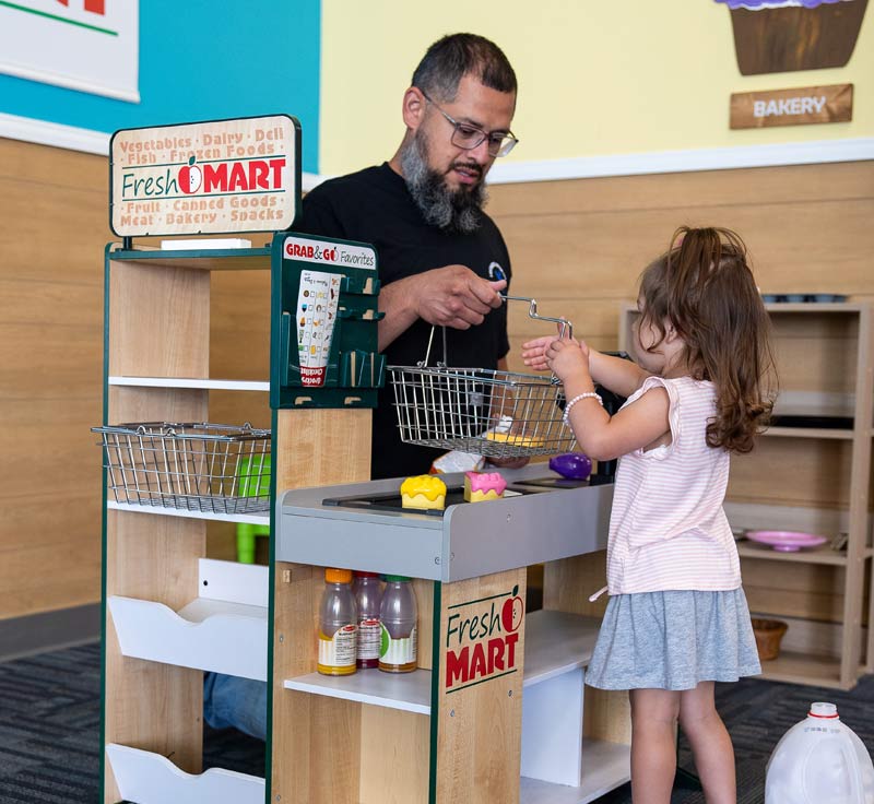 A therapist with short black hair, glasses, and a black and grey beard kneeling at a toy checkout as a lighthouse learner hands him a shopping basket.