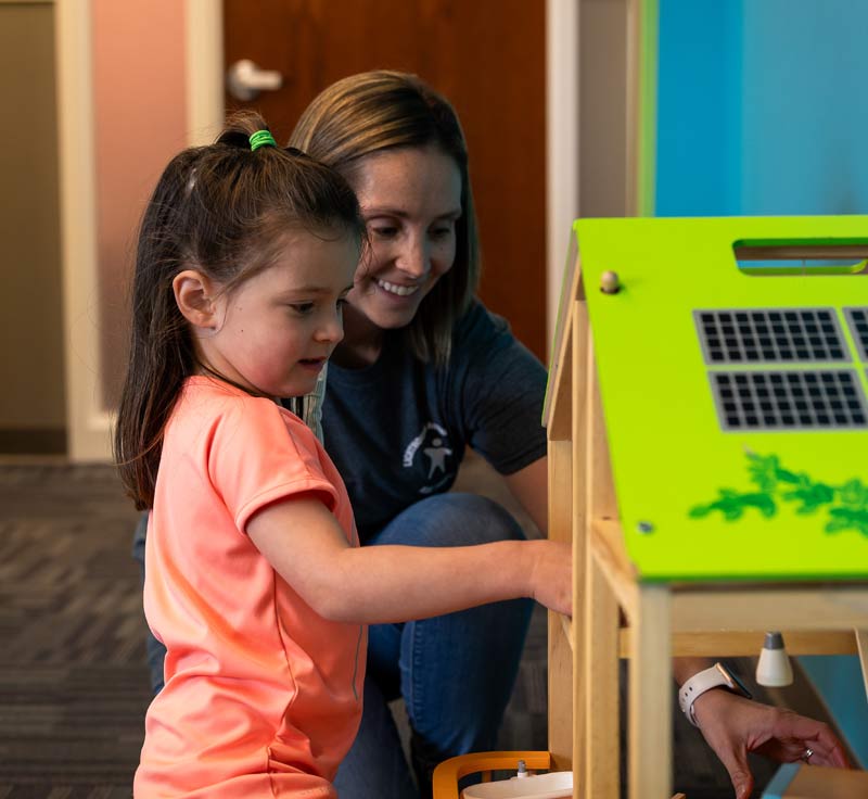 A Lighthouse Autism Center staff member smiling next to a child playing with a wooden doll's house with a green roof