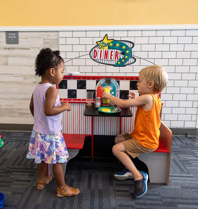 Two children sat and stood playing at toy food diner