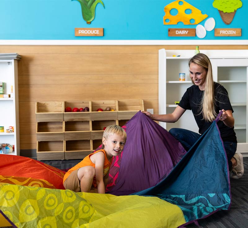 A Lighthouse Autism Center staff member with blonde hair holding a multi-colored parachute and a child crouched inside