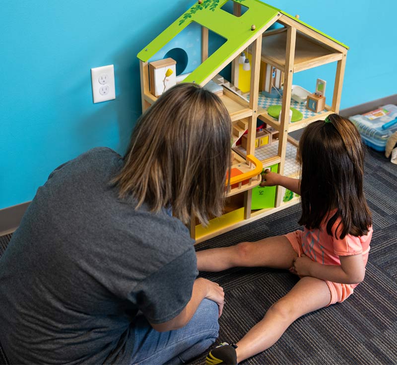 A woman and a child sat on the carpet playing with a wooden green and yellow doll's house in front of a blue wall.