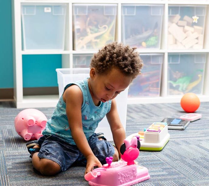 A child wearing a blue t-shirt and denim shorts knelt down in front of clear plastic boxes playing with a pink toy cash register
