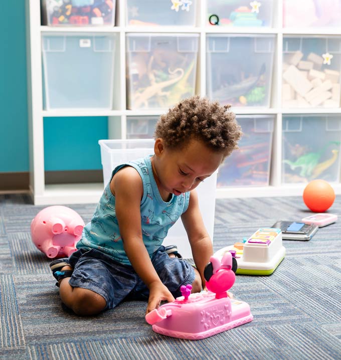 A child knelt on the floor in front of boxes filled with toys playing with a pink toy cash register.