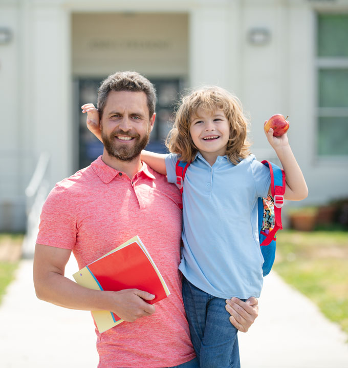 A man with gray hair and a beard wearing a red shirt smiling stood holding his son wearing a blue shirt and backpack