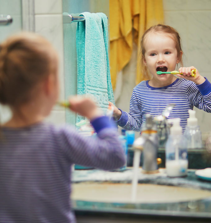 A child stood looking into a mirror by the sink in a bathroom with a tap running brushing her teeth