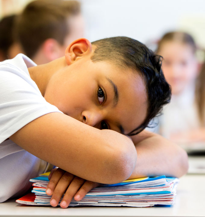 A child with black hair who has his arms crossed and head resting on a stack of books on a desk at school