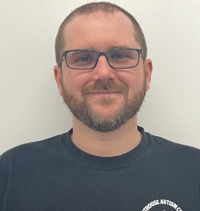 A man with glasses, short hair and a short beard wearing a black Lighthouse Autism Center shirt in front of a pale wall.