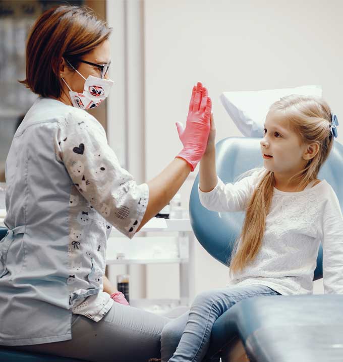 A doctor wearing a white uniform with a face mask high-fiving a child with blond hair sat on a medical chair.