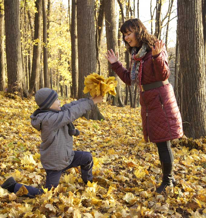 A woman stood up wearing a long brown coat in a wooded area at fall with a child knelt down holding a bunch of yellow leaves.