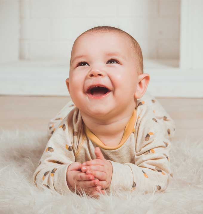 A baby in a beige outfit laying on a rug looking up smiling.