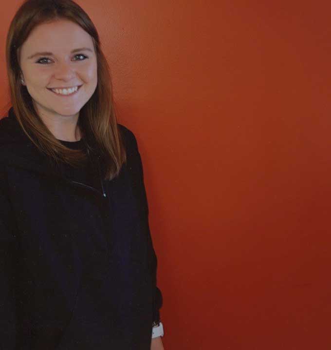 A Lighthouse Autism Center employee named Amanda in a black jumper smiling stood in front of an orange wall