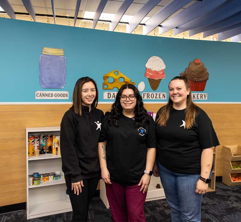 Three female Lighthouse Autism Center employees wearing black t-shirts smiling in front of a blue wall