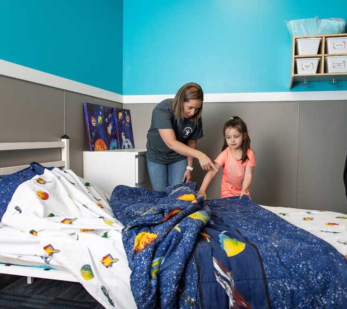 A Lighthouse Autism Center staff member helping a child make a bed pointing at the space themed comforter covers