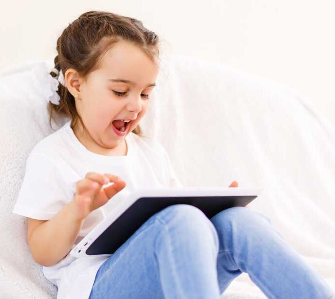 A child sat on a white sofa wearing a white t-shirt and jeans holding a tablet with a happy facial expression