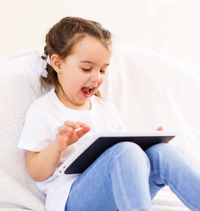 A child wearing a white t-shirt sat on a sofa smiling at a tablet